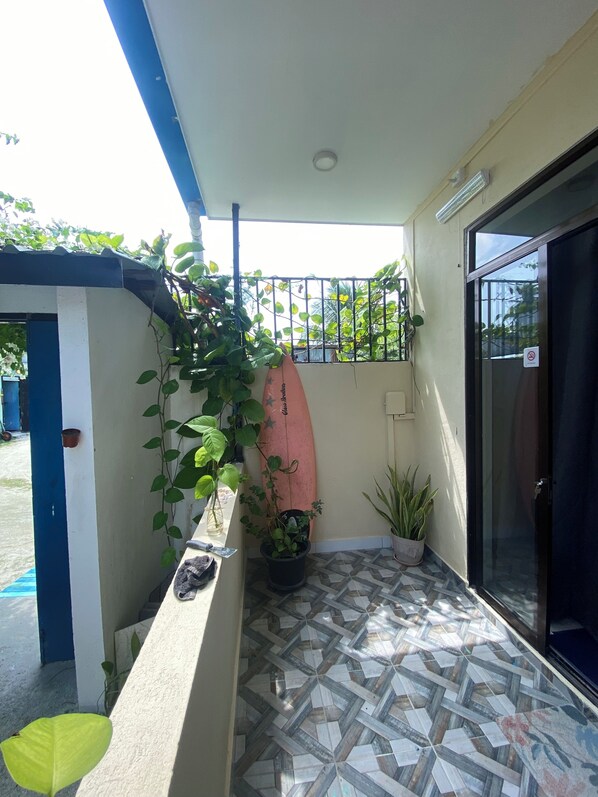 A small balcony with a tiled floor, a potted plant, a surfboard, and a door leading to a covered area. - Maavaharu K.Himmafushi  (Himmafushi)