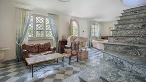 A room with a checkered floor, a brown leather sofa, a wooden chair, a marble staircase, and a view through a window with a lattice design.