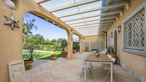 A covered patio with a table and chairs, potted plants, and a view of a landscaped garden.