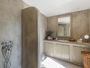 Bathroom features a large, curved shower enclosure with handheld showerhead and flexible hose, a vanity with large mirror and sink, and a small stool.