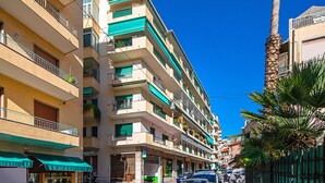 Un immeuble à plusieurs étages doté de balcons, une rue en face et un palmier sur la droite.