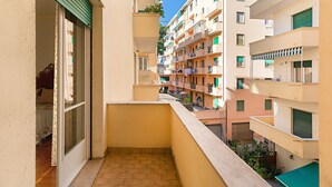 Un balcon étroit offrant une vue sur les immeubles voisins, tous dotés de plusieurs étages et agrémentés de balcons où l’on peut voir des plantes et du linge séchant.