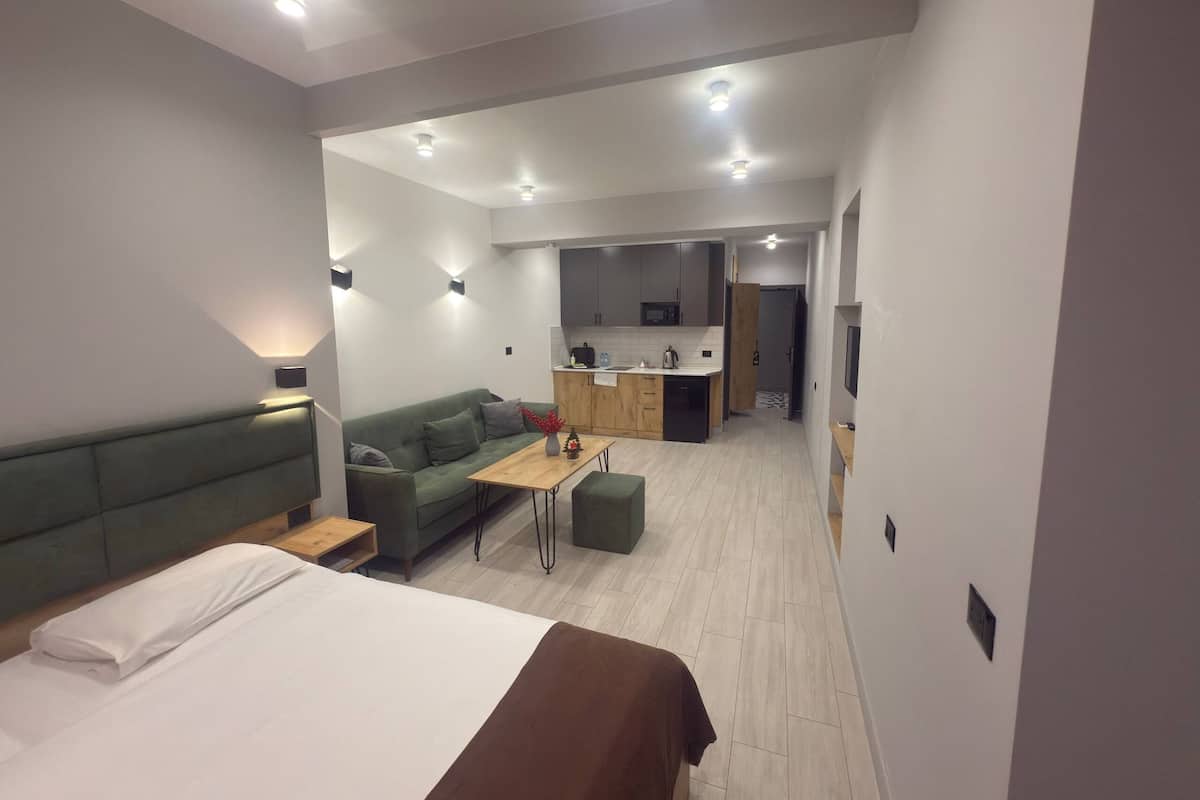 A modern hotel room with a bed, a green sofa, a small table, and a kitchenette in the background.