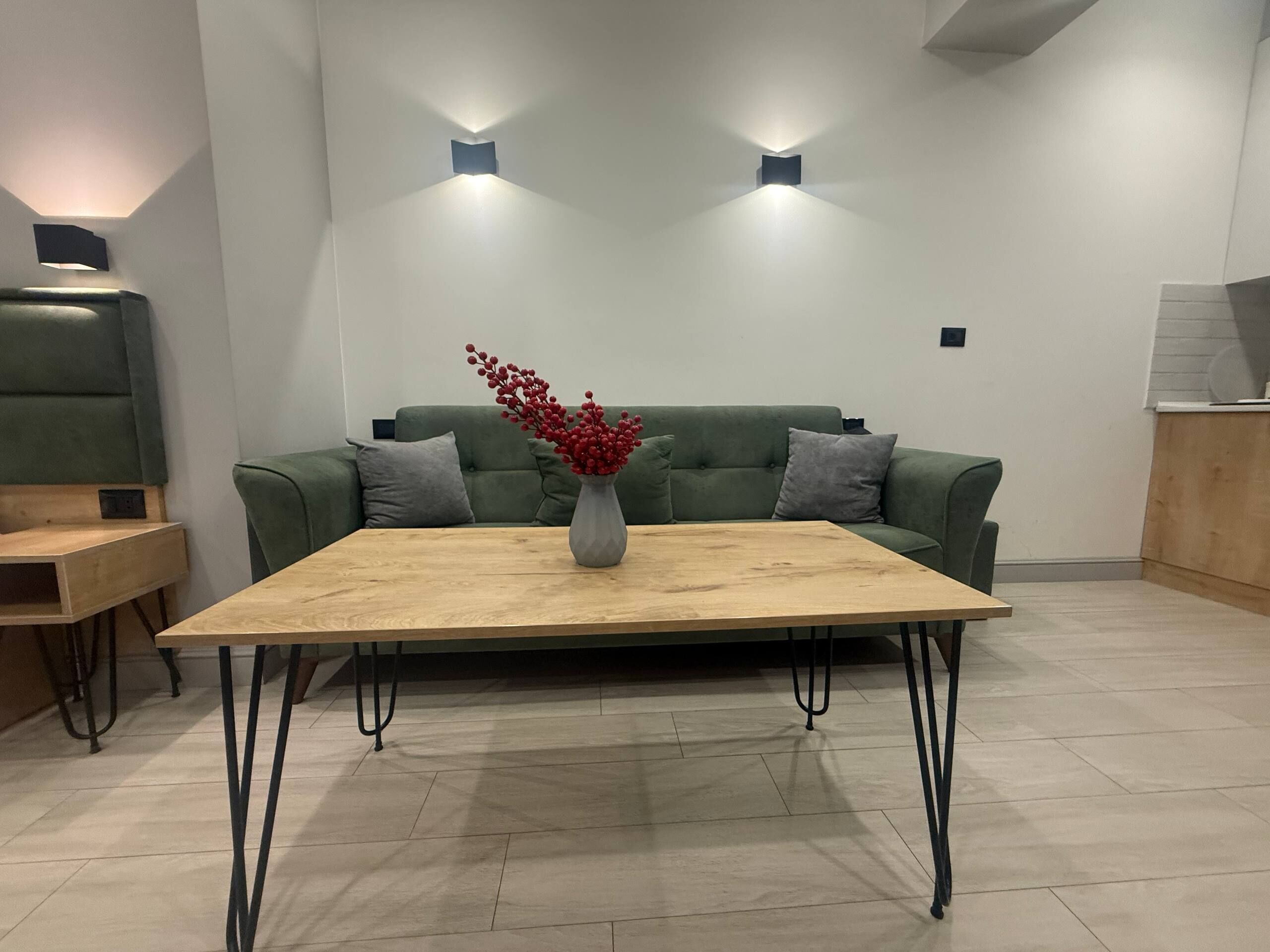 Un salón moderno con un sofá verde, una mesa de centro de madera y un jarrón con flores rojas. Hay dos luces fijadas a la pared sobre el sofá.