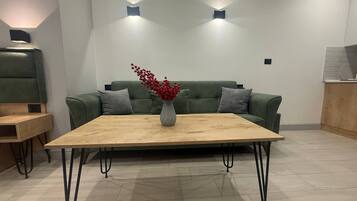 Un salón moderno con un sofá verde, una mesa de centro de madera y un jarrón con flores rojas. Hay dos luces fijadas a la pared sobre el sofá.