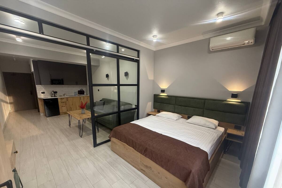 A modern bedroom with a large bed, a desk, and a glass partition separating the bedroom from a kitchenette.