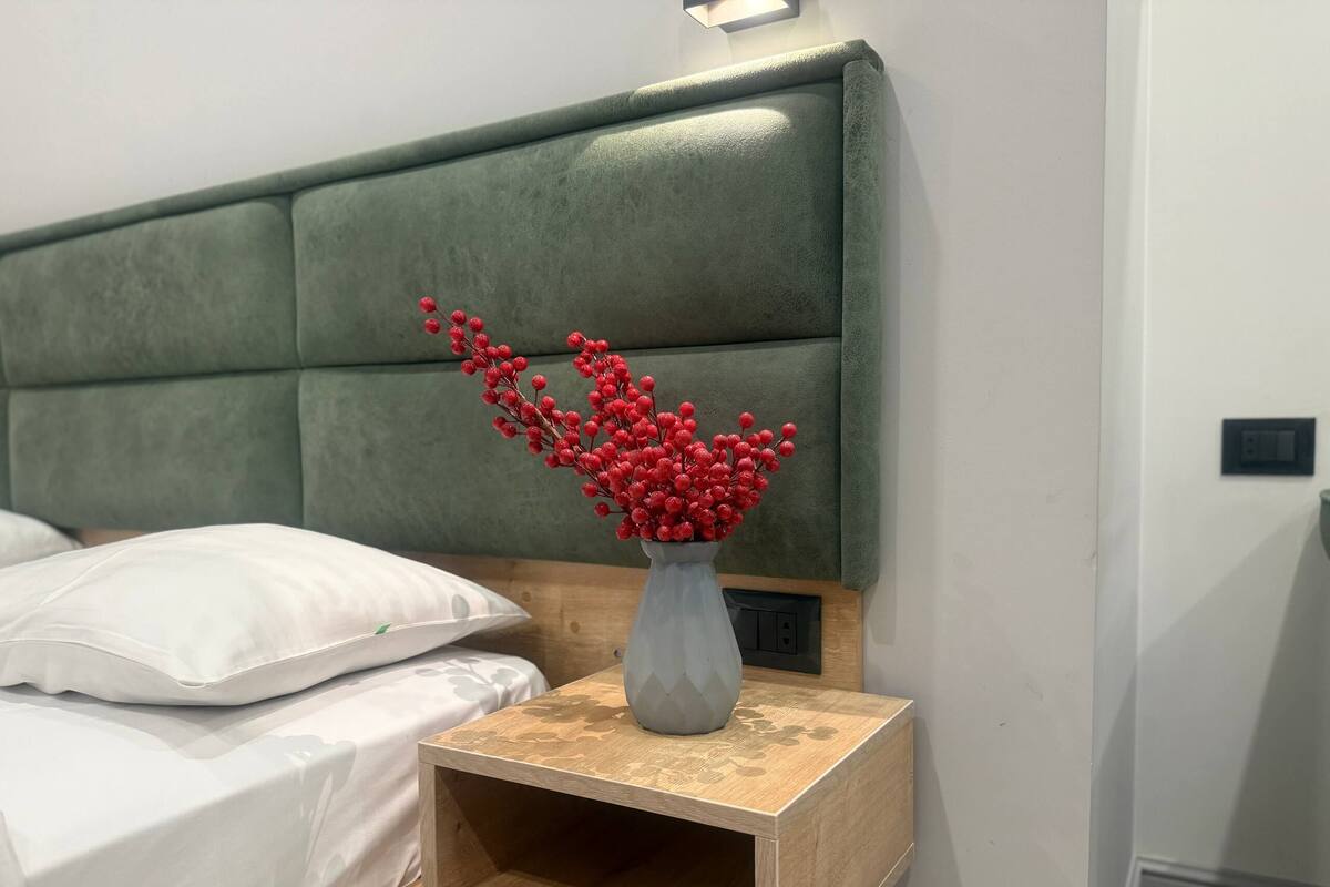 A neatly made bed with white linens, a wooden nightstand with a vase of red berries, and a green upholstered headboard.