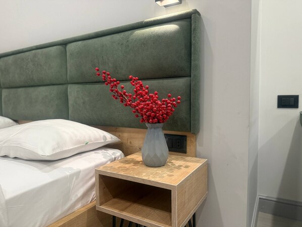 Ein ordentlich bezogenes Bett mit weißen Leinen, ein Nachttisch aus Holz mit einer Vase voller roter Beeren und ein gepolsterter, grüner Kopfteil.