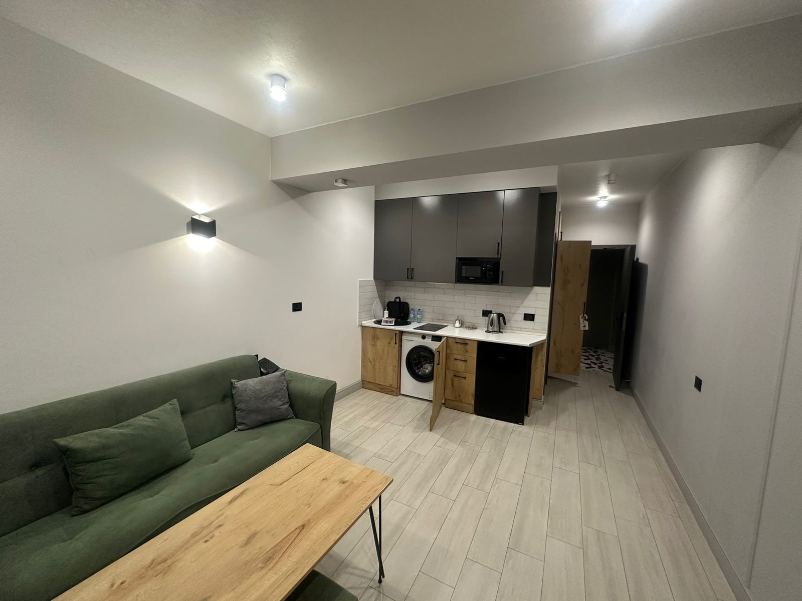 Een compacte woonruimte met een groene bank, een houten salontafel, een wasmachine en een kitchenette met een magnetron.