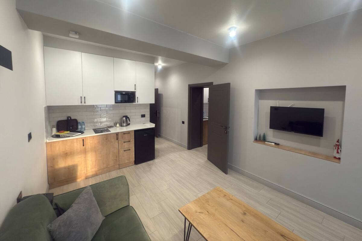 A modern kitchen with white cabinets, a microwave, and a coffee maker. There is a green sofa and a wooden table in the living area. A TV is mounted on the wall.