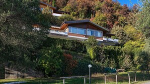 A modern house with a balcony, surrounded by lush greenery and trees with autumn foliage.