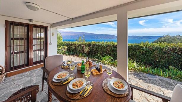 A dining table set with plates of food, glasses, and bottles, overlooking a scenic view of the sea and hills.