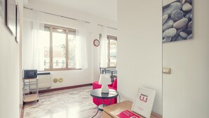 Quarto de hotel com cama, escrivaninha com luminária, cadeira, mesinha e janela com cortinas.