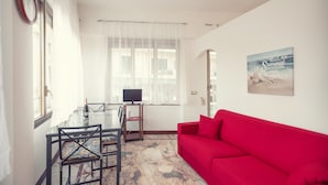 Uma sala de estar com um sofá vermelho, uma mesa de jantar de vidro com cadeiras, uma televisão sobre um móvel e uma janela com cortinas leves.