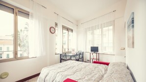 Quarto com cama, escrivaninha e cadeira. Há uma janela com cortinas, um relógio na parede e um pôster na parede.