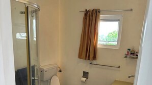 A compact bathroom with a glass shower enclosure, a toilet, a sink, and a small window with curtains.