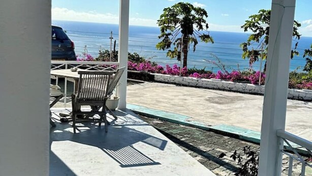 A covered outdoor area with a view of the ocean, a table, a chair, and potted plants.