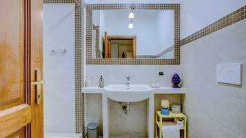 A bathroom with a white sink, mirror, and toilet. There is a small trash can and a shelf with toiletries.