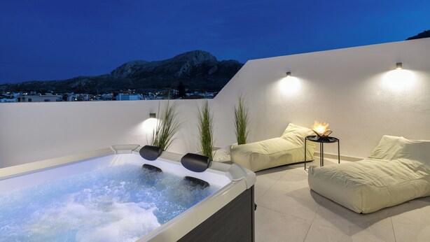 A modern outdoor hot tub area with a view of a mountain, a white wall, and a sofa with cushions.