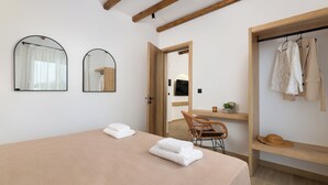 A bedroom with a bed, two mirrors, a wooden chair, a wooden desk, and a wardrobe with hanging clothes.