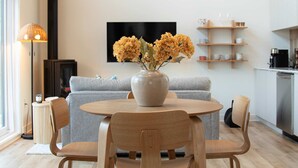 A modern living room with a round wooden table, two chairs, a grey sofa, a vase with flowers, a flat-screen TV, and a kitchen area with white cabinets and a coffee machine.