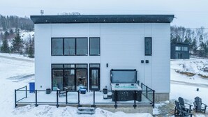A modern two-story building with large windows, a covered outdoor area with a hot tub, and a snowy landscape.