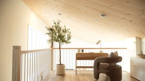 A modern living room with a wooden ceiling, a potted tree, a sofa, and a desk with a lamp.