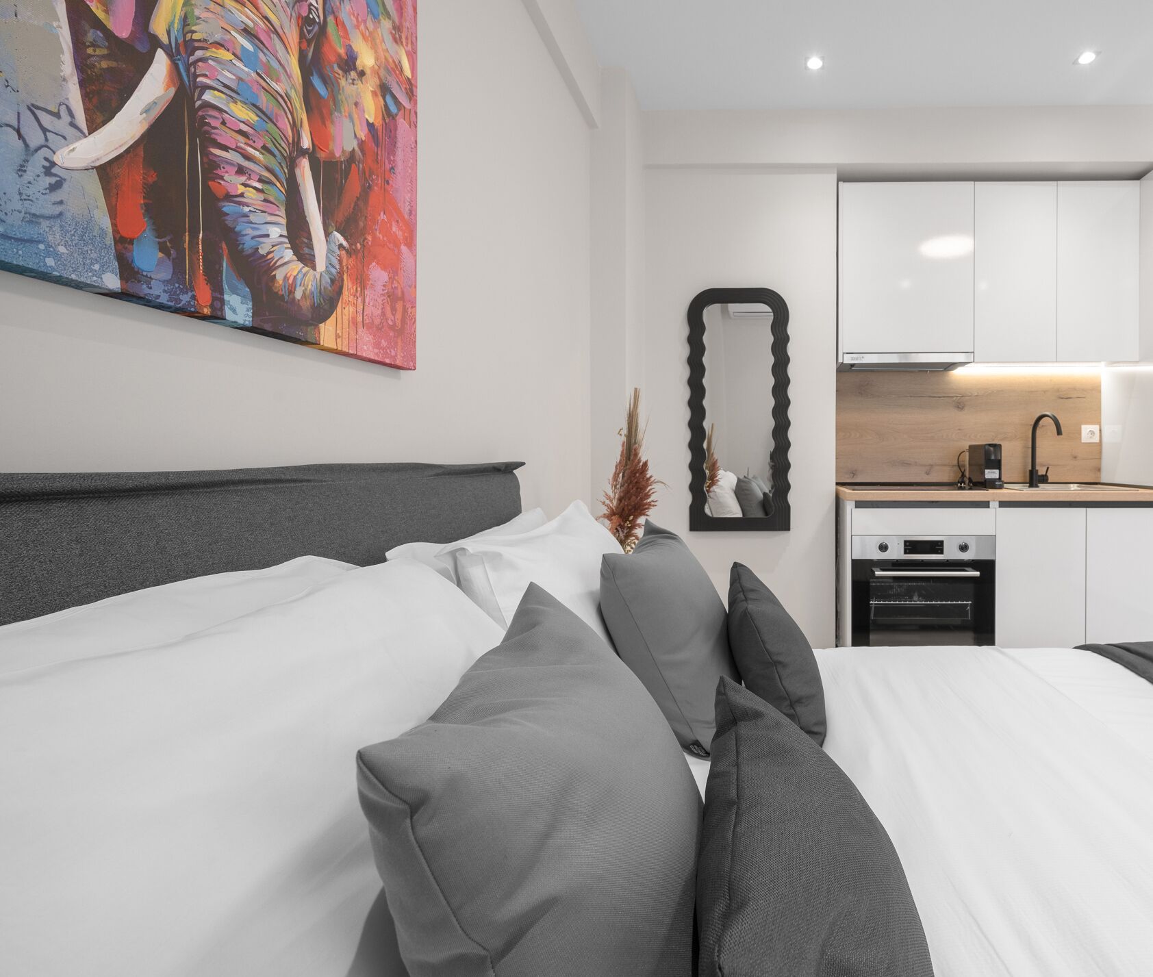 A modern hotel room with a large bed, a small kitchenette, and a colorful elephant painting on the wall.