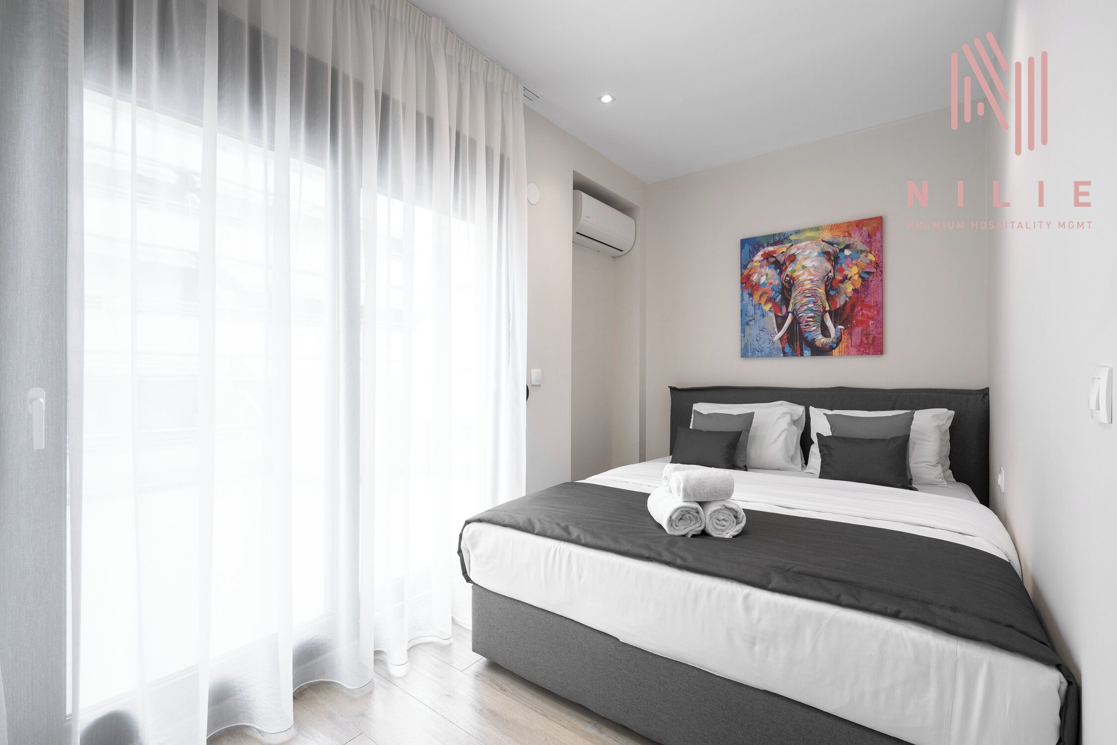 A modern hotel room with a large bed, white curtains, an air conditioning unit, and a colorful elephant painting on the wall.