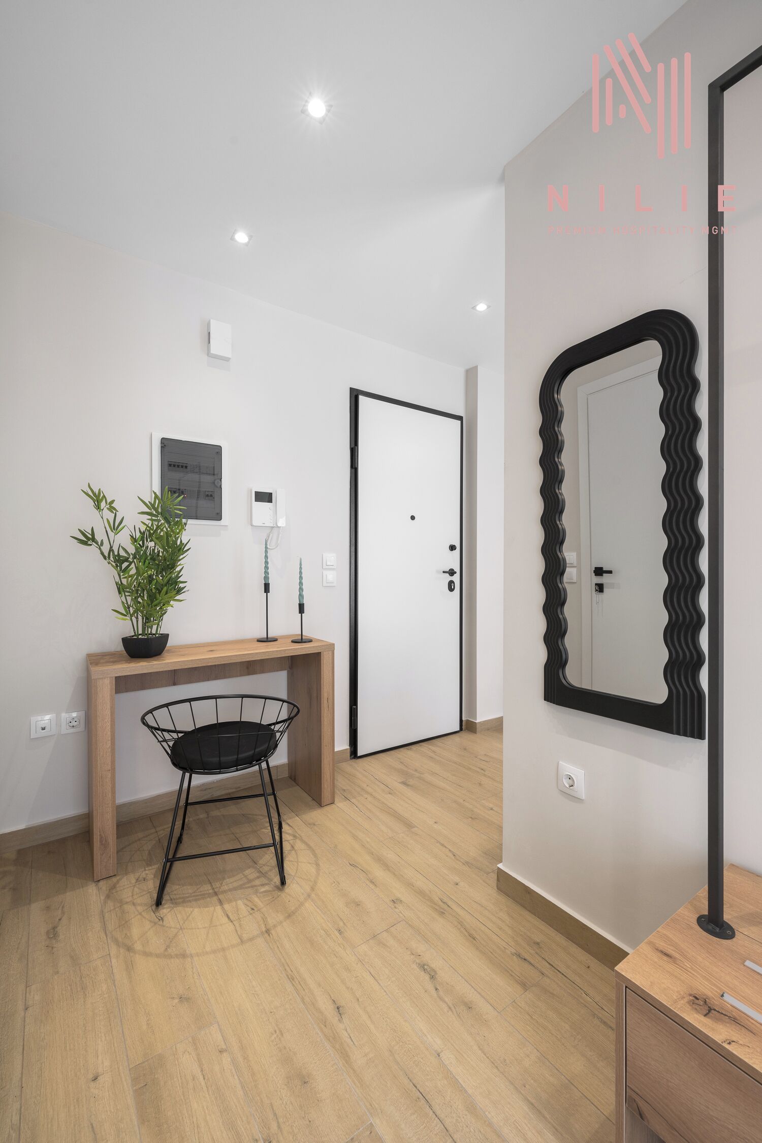 A modern room with a wooden console table, a black wire chair, a potted plant, a wall-mounted mirror, and a white door.