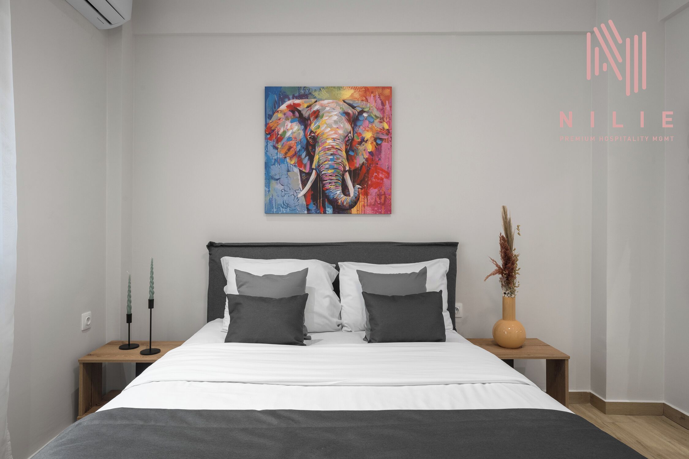 Ein modernes Schlafzimmer mit einem großen Bett, zwei Nachttischen, einem Wandbild eines Elefanten und dem Logo von 'NILIE Premium Hospitality Management'.