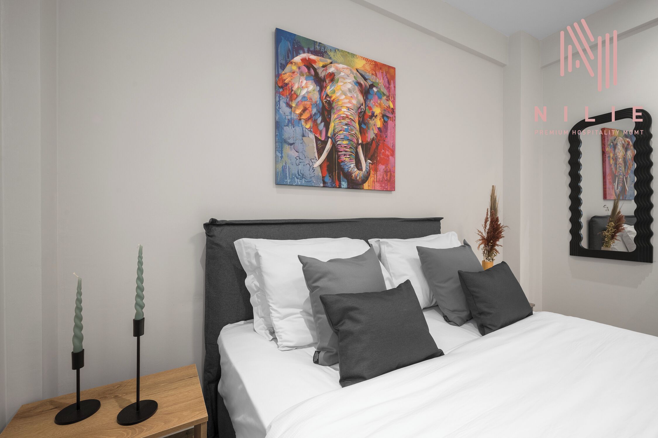 A modern bedroom with a large bed, a colorful elephant painting, and a black mirror on the wall.