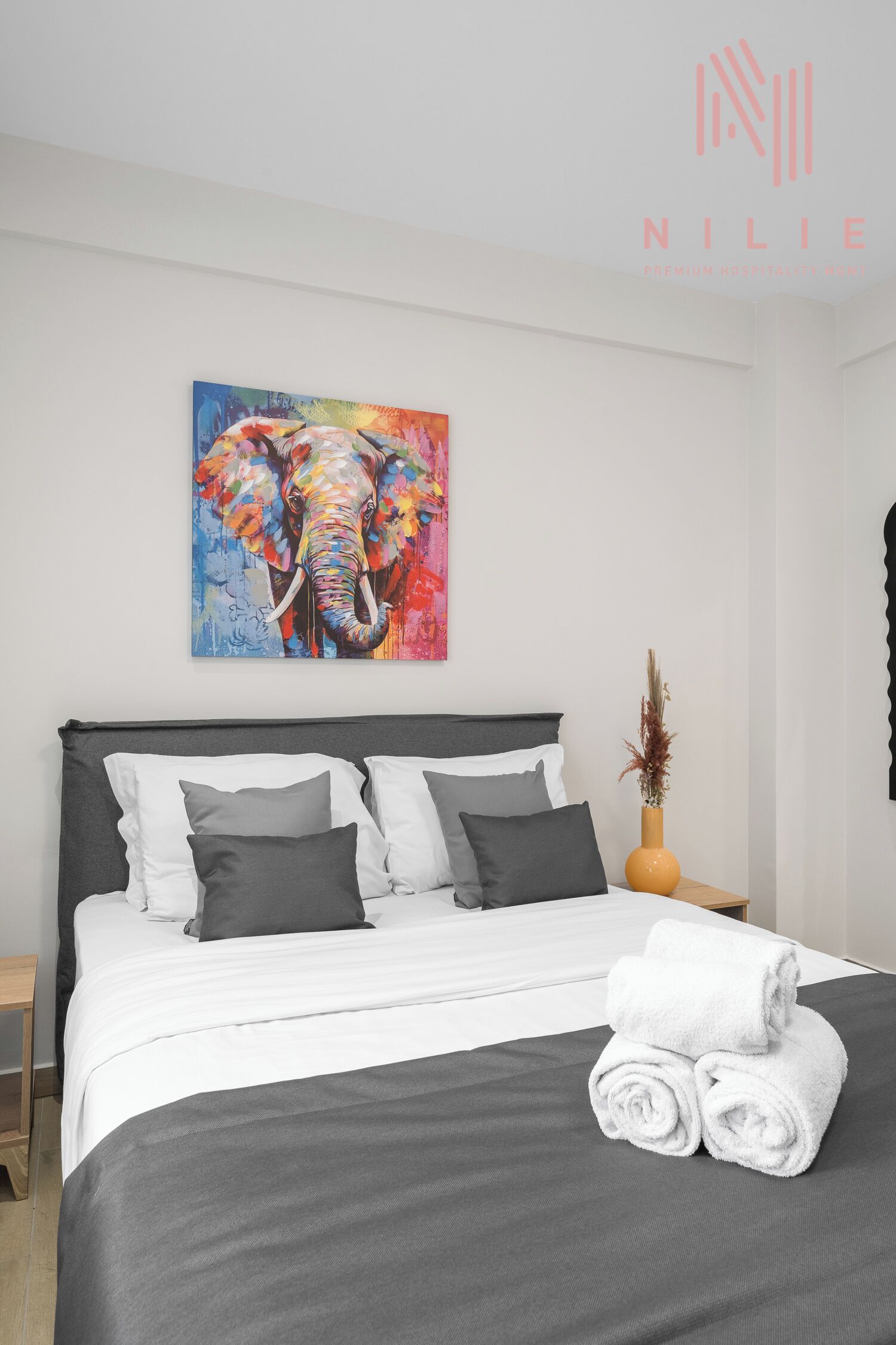 Un lit bien fait, avec une literie blanche et grise, un tableau coloré représentant un éléphant accroché au mur, et un vase contenant des plantes séchées sur la table de chevet.