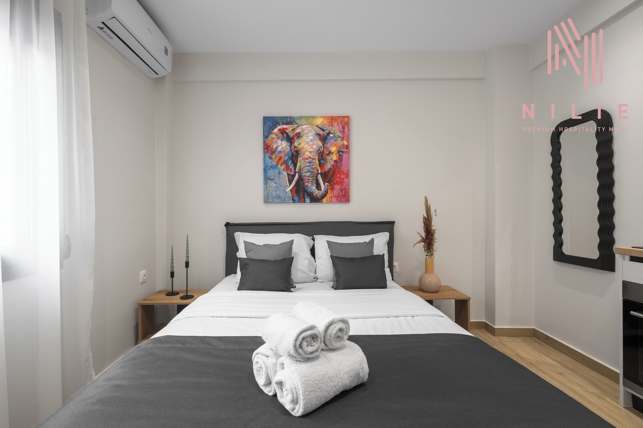 Una habitación de hotel moderna con una cama grande, un cuadro de un elefante, un espejo y una mesita de noche con un florero.