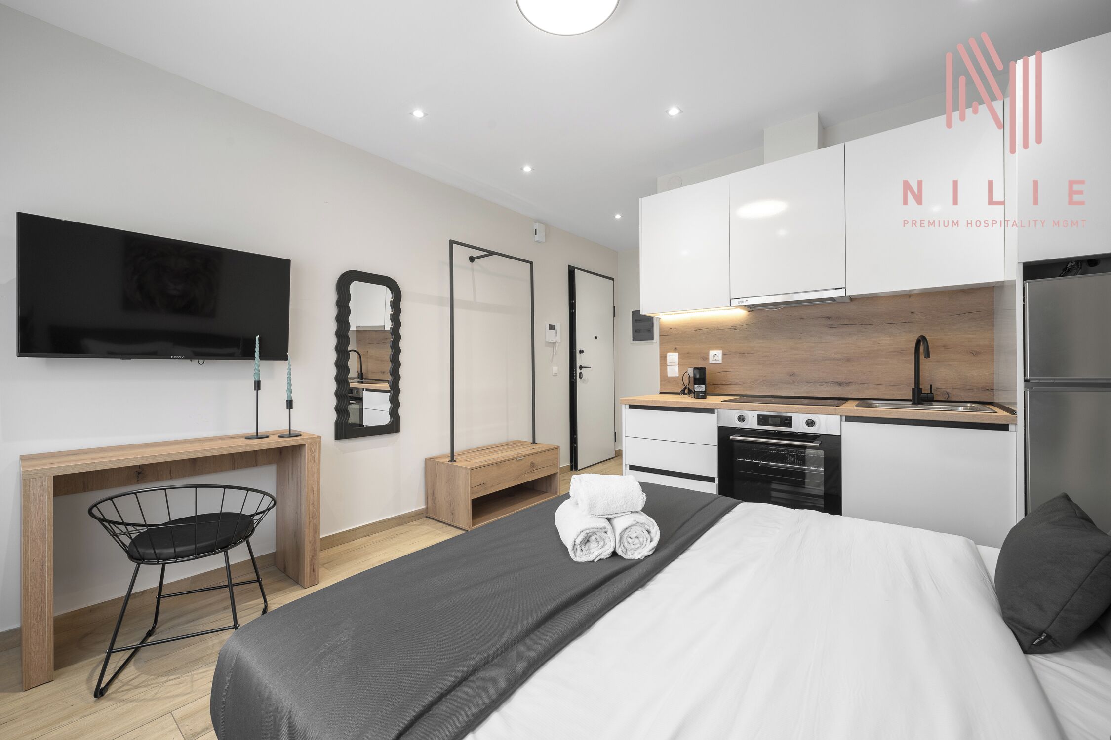 A modern hotel room with a bed, a desk, a chair, a TV, a mirror, a sink, and a kitchen area.