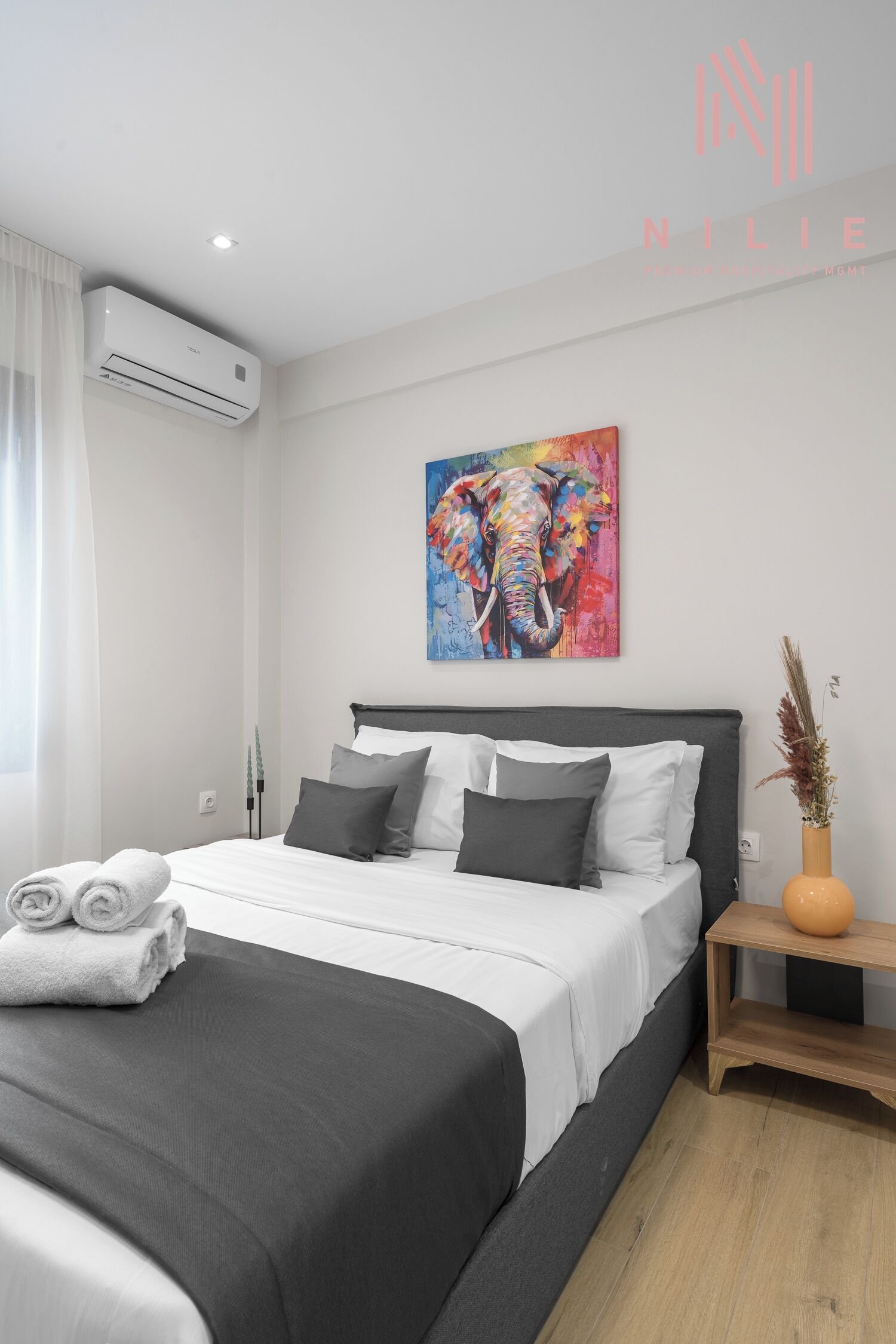 A modern bedroom with a large bed, a painting of an elephant, and a vase with dried plants.