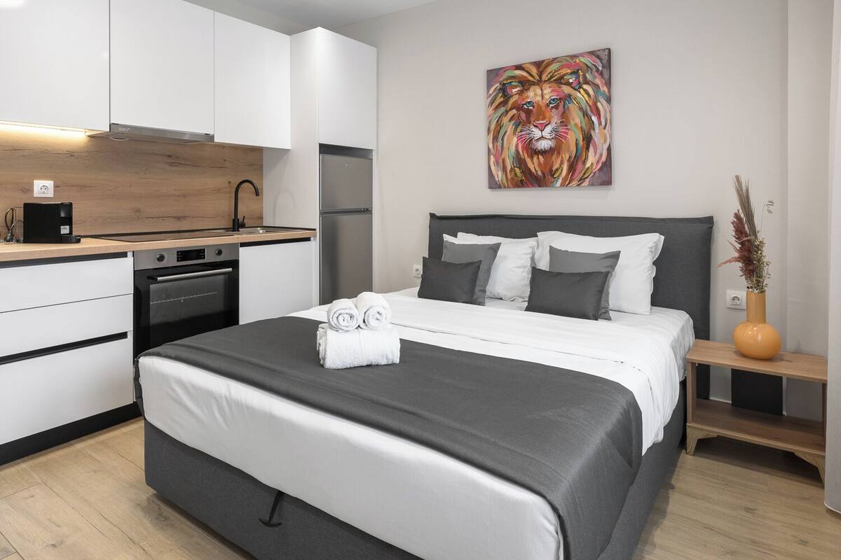 A modern bedroom with a large bed, a bedside table with a vase, and a kitchen area with white cabinets and a wooden countertop.