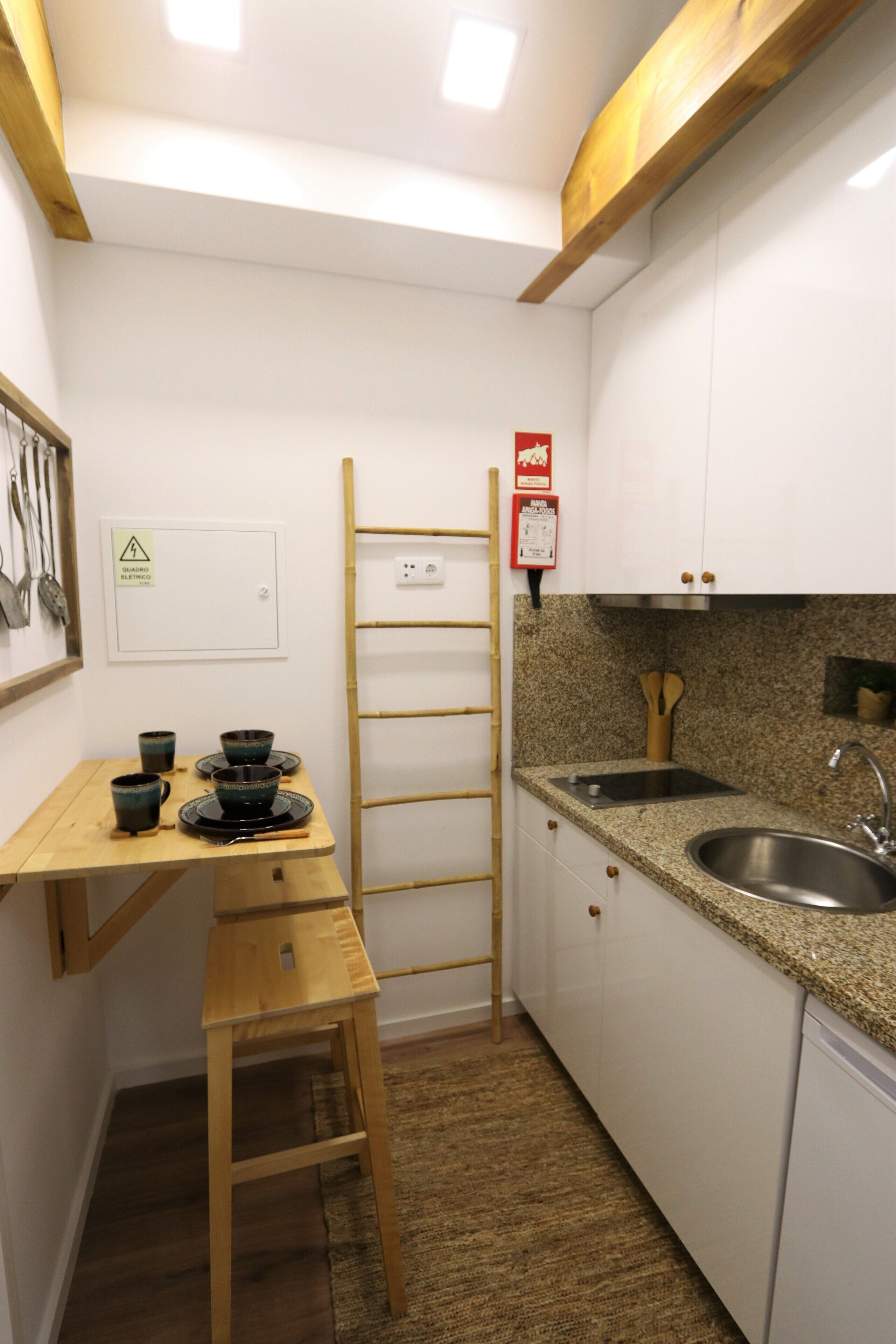 Une cuisine compacte avec des meubles blancs, un plan de travail en granit, un évier en acier inoxydable, une échelle en bois et une petite table avec des tasses.