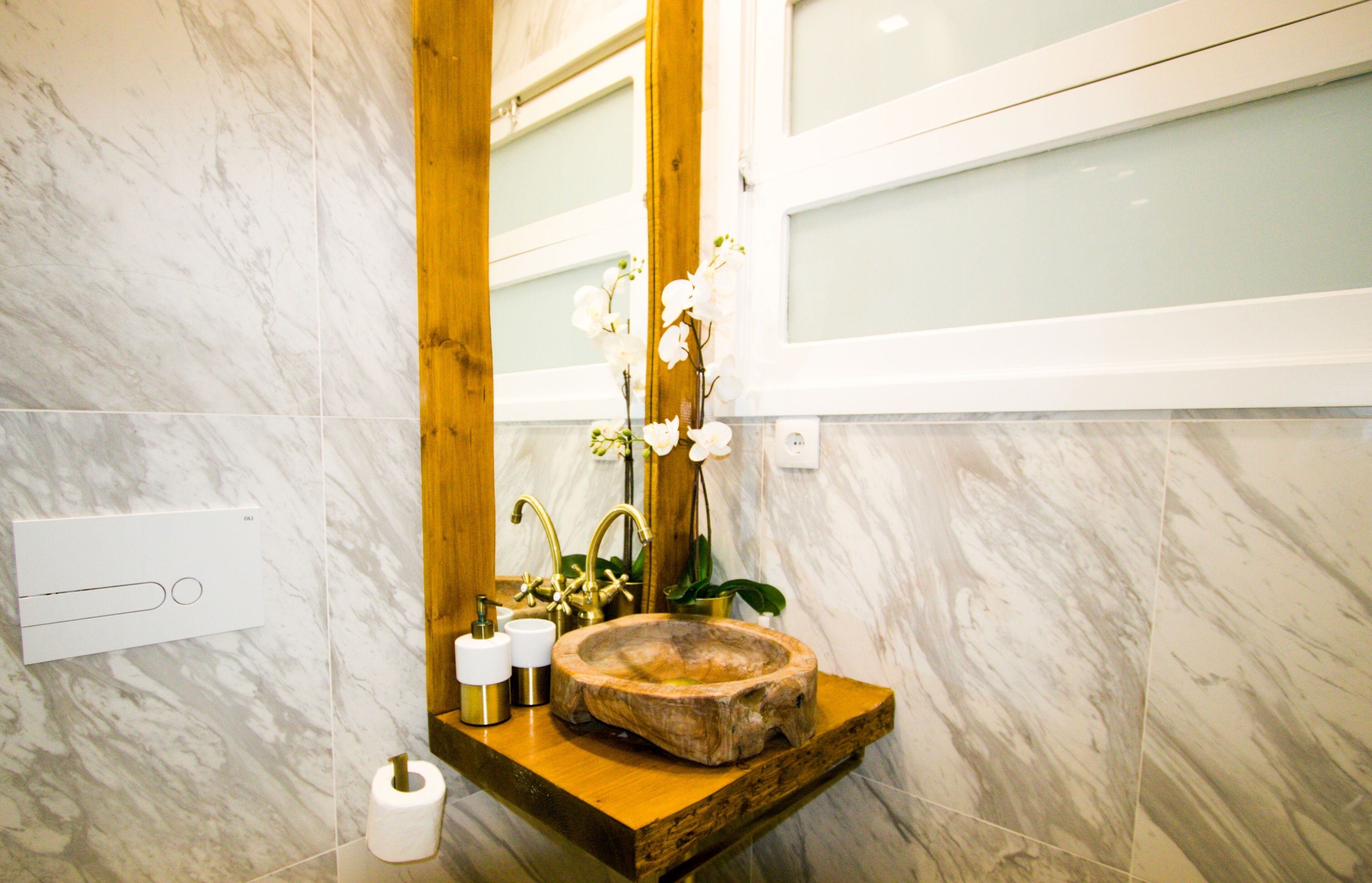 A bathroom with a marble wall, wooden vanity, a sink with a wooden bowl, a mirror, and a toilet paper holder.