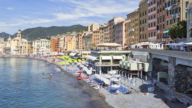 A coastal town with a beach lined with umbrellas and sun loungers, buildings with balconies, and a clear sky.