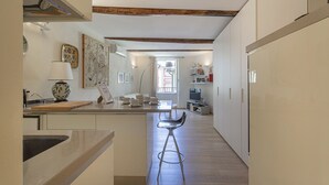 A narrow kitchen with a central island, wooden beams on the ceiling, and a view of the outside through a window.