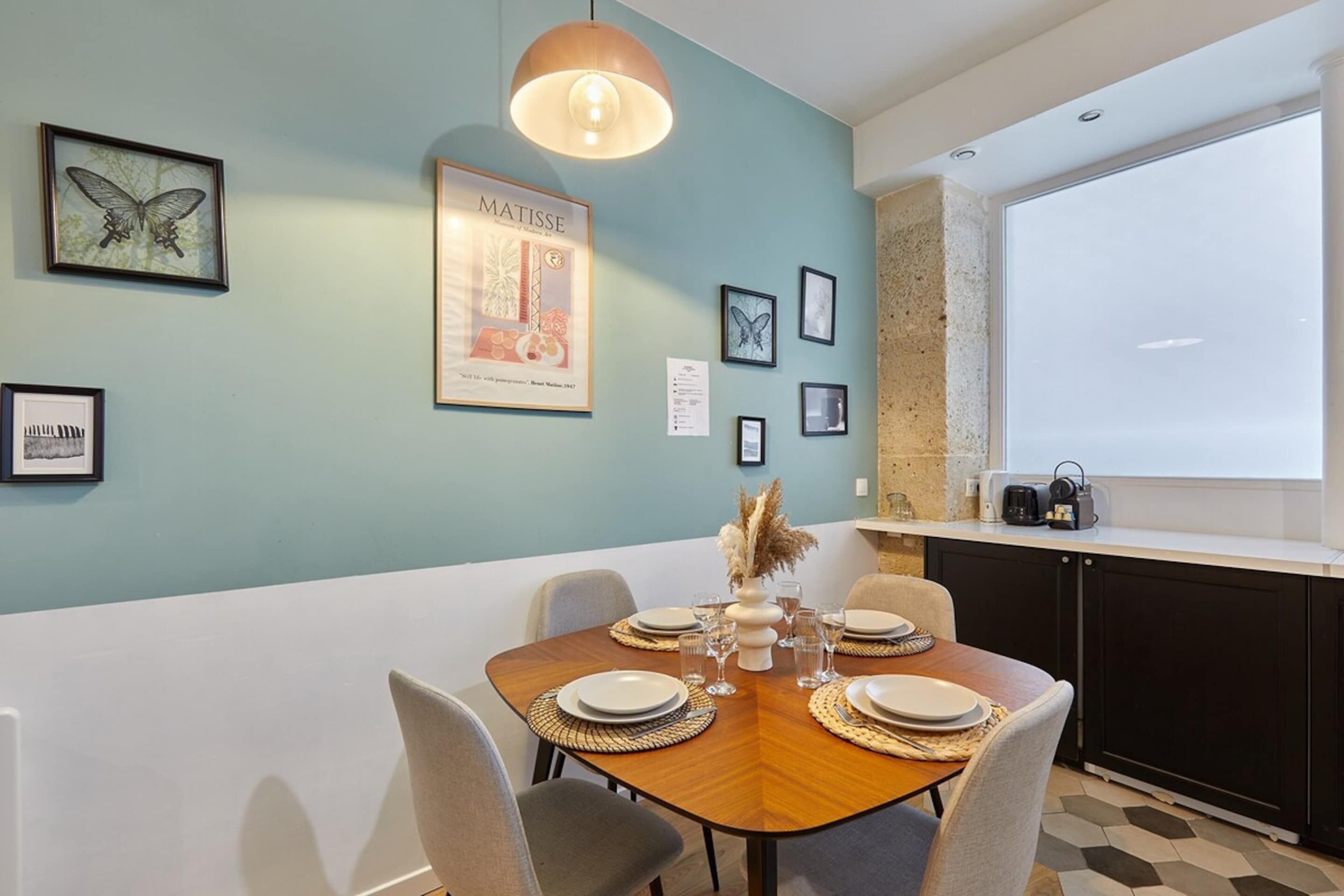 A dining area with a wooden table set for four, surrounded by chairs. There are framed artworks on the walls, including a prominent Matisse poster.