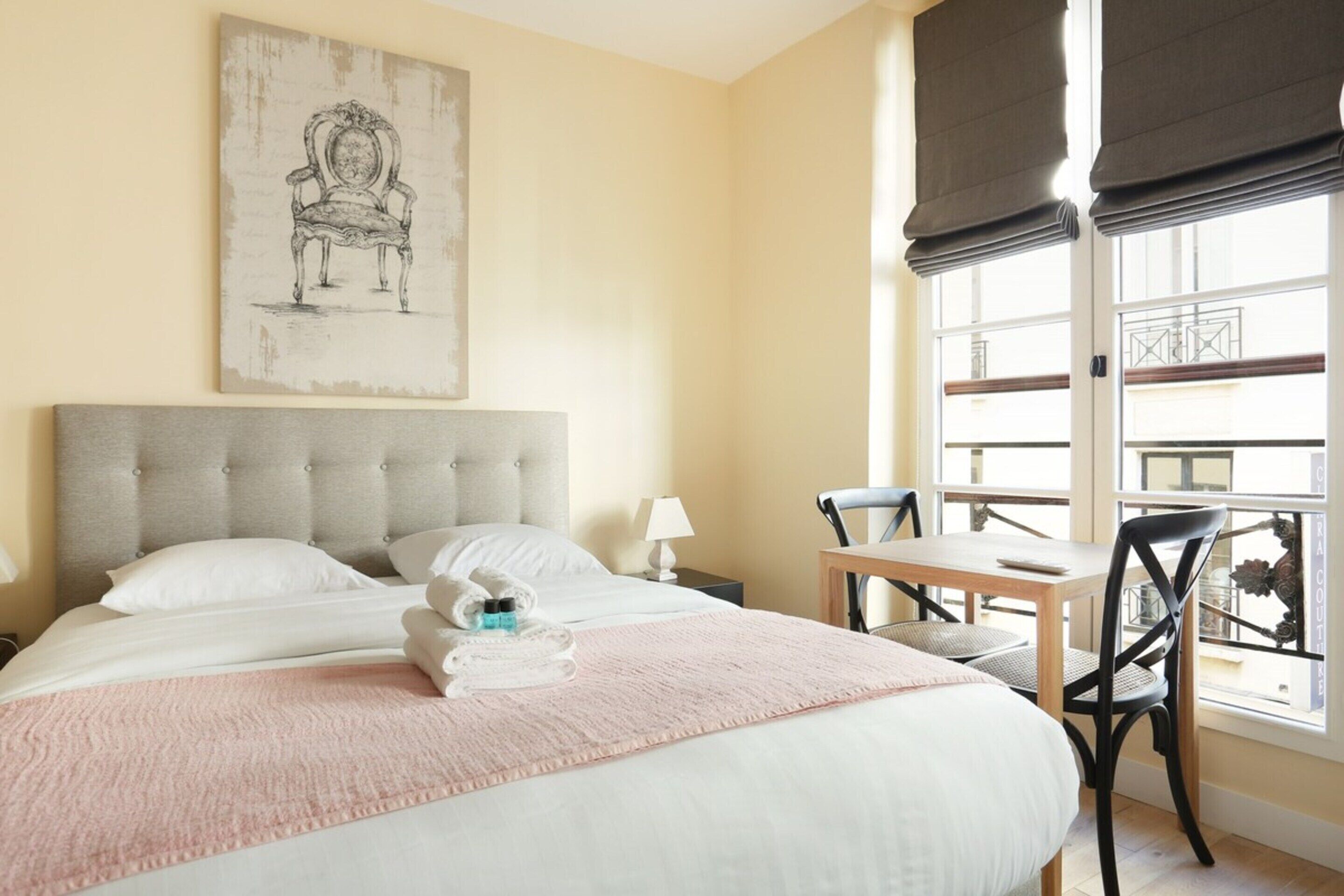 A hotel room with a large bed, a desk, and a chair. There is a window with a view of the outside.