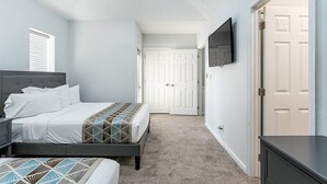 Un dormitorio con cama, mesitas de noche, televisor montado en la pared y una puerta que da a otra habitación.