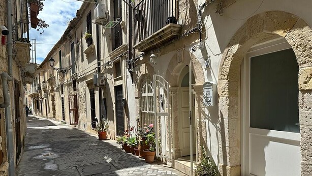 Una estrecha calle adoquinada flanqueada por edificios históricos, plantas en macetas y una puerta blanca.