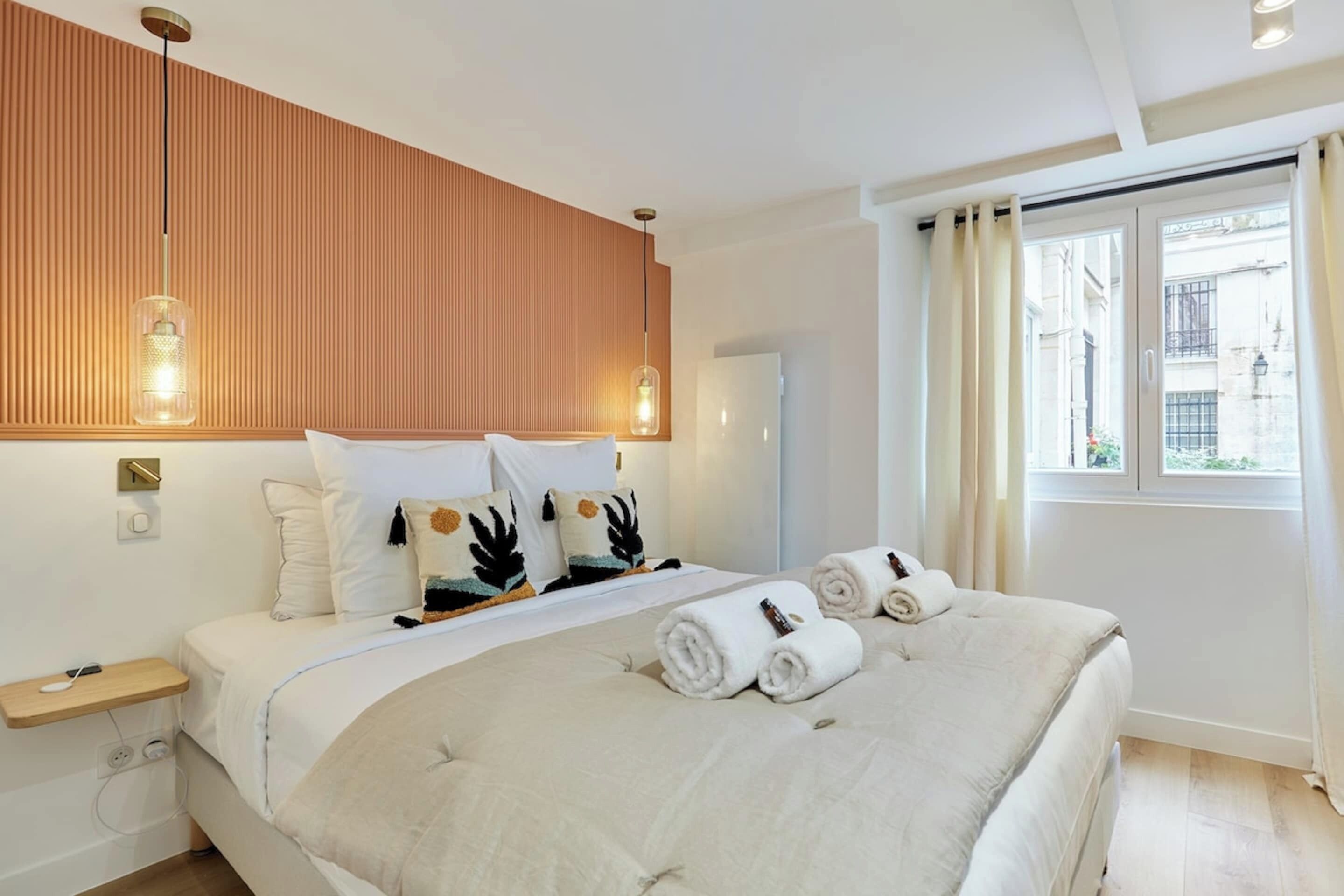Una habitación de hotel moderna con una cama grande, un panel de pared de madera, dos lámparas colgantes, una ventana con cortinas y una mesita de noche pequeña.