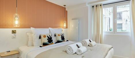 Una habitación de hotel moderna con una cama grande, un panel de pared de madera, dos lámparas colgantes, una ventana con cortinas y una mesita de noche pequeña.