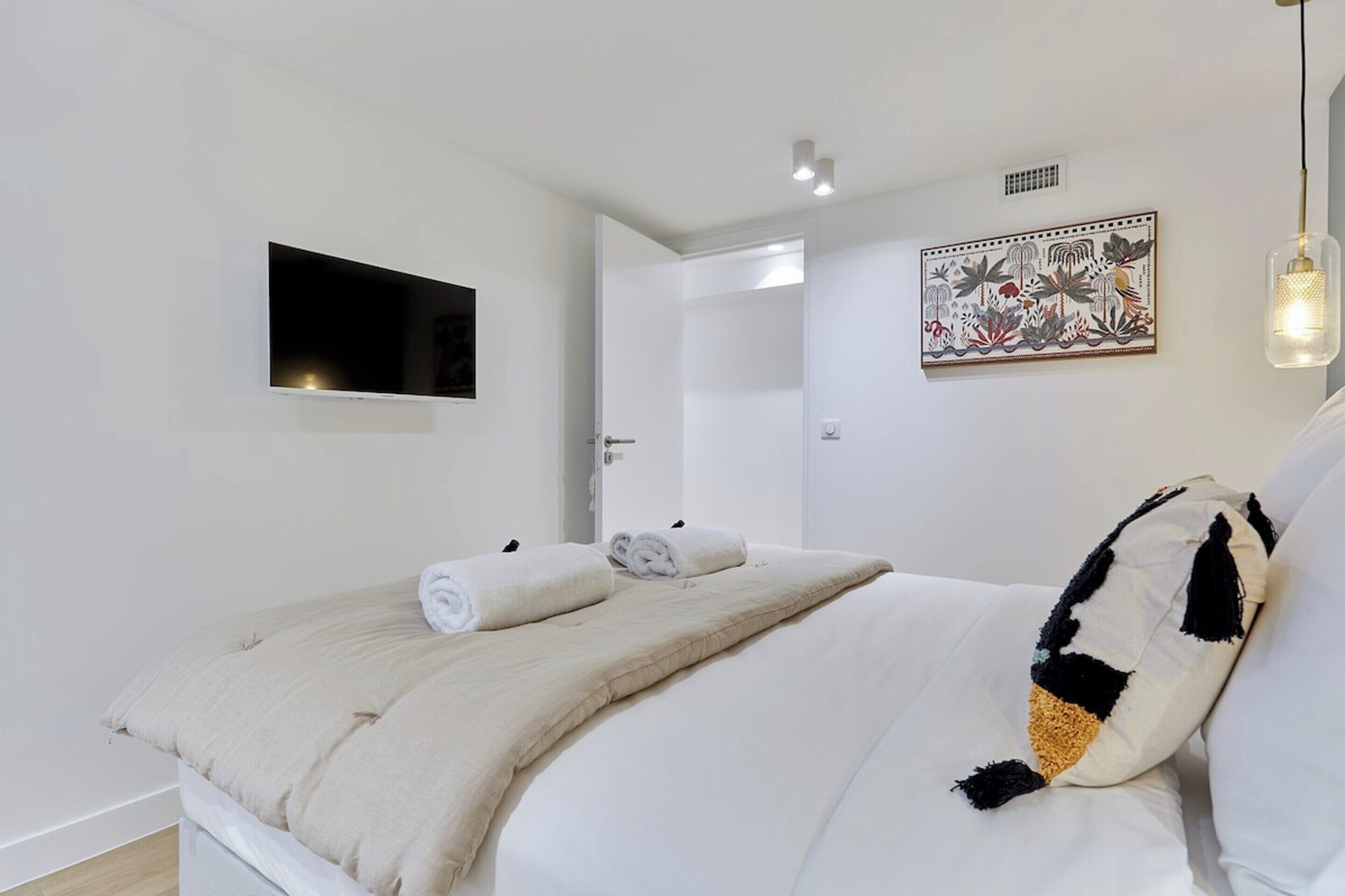 A neatly arranged bedroom with a bed, a flat-screen TV mounted on the wall, a decorative wall piece, and a hanging light fixture.
