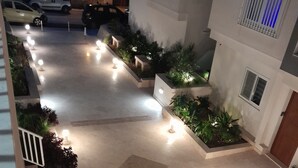 En gårdsplads med fliset gulv, omgivet af plantekasser med grønne planter, og oplyst af flere lamper.