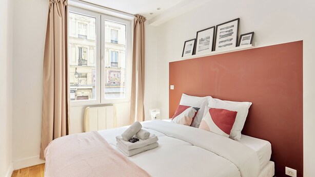 Une chambre d’hôtel avec un grand lit, des rideaux beiges, une fenêtre donnant sur des immeubles et un mur d’accent rouge.